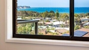 A room with a large window overlooking a coastal area, a bed with a patterned bedspread, and a visible railing. - Seaside Escape (Bega Valley)