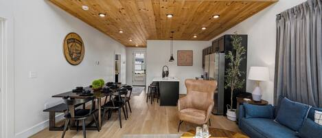 A modern living room with a dining area, a wooden ceiling, and a view of the kitchen.