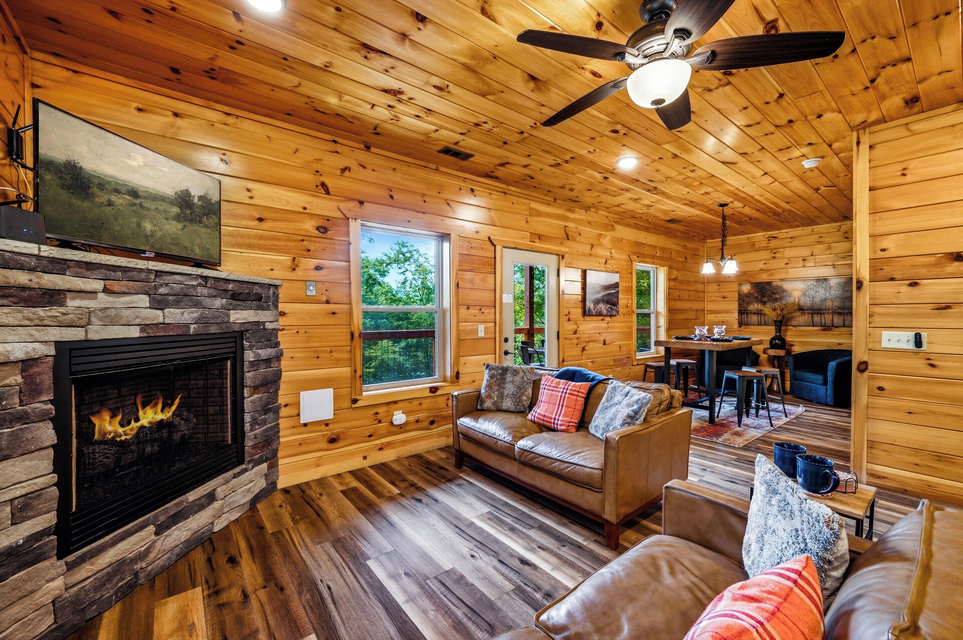 A log cabin interior with a stone fireplace, wooden floors, a ceiling fan, and a dining area.