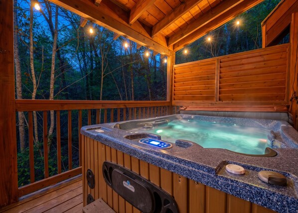 Una terraza de madera con un jacuzzi, rodeada de árboles y luces colgantes.