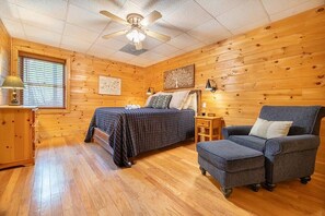 Ein Zimmer mit Holzwänden, einem Bett, einem Sessel, einem Nachttisch und einem Deckenventilator.