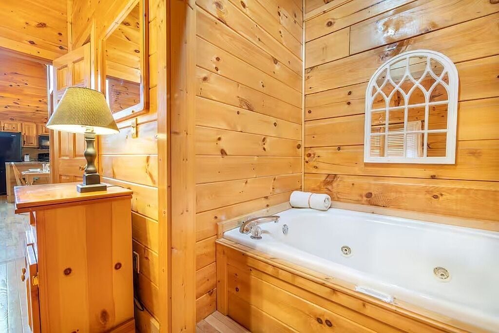 Interior de una cabaña de madera con una bañera de madera, una lámpara y una ventana con una celosía decorativa.