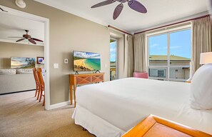 Ein Hotelzimmer mit Bett, Fernseher, Deckenventilator, einem Fenster mit Vorhängen und Blick auf den Strand.
