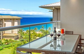 A balcony with a glass table, two wine glasses, and bottles, overlooking a beach and ocean. - 7th-Floor Honua Kai 1BR with Ocean and Mountain Views (Lahaina)
