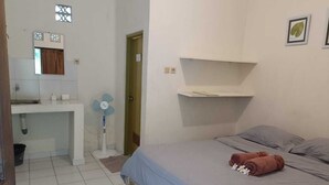 A bedroom with a bed, a bedside table, a wall-mounted shelf, and a bathroom with a sink and a fan.