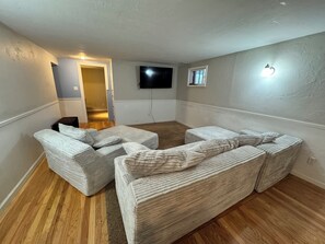 A room with a large sectional sofa, a flat-screen TV mounted on the wall, and a small window.
