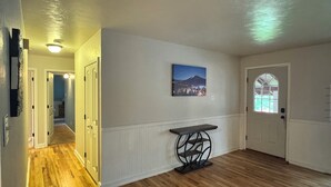 Un couloir avec un plancher en bois, des murs blancs, une porte avec une fenêtre et une petite table sur laquelle est posée une image accrochée au mur.