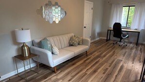 Ein Zimmer mit Holzboden, einem weißen Sofa mit gemusterten Kissen, einem kleinen Beistelltischchen mit Lampe und einem Schreibtisch mit Stuhl.