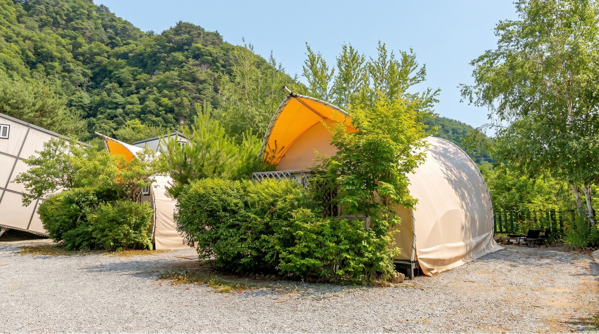 Un camping con tiendas de campaña y una estructura grande cubierta, rodeado de árboles y una colina al fondo.