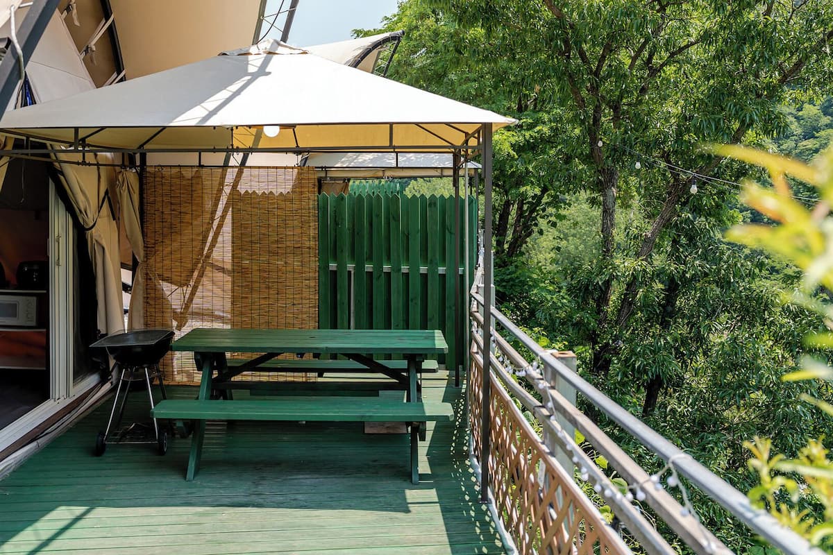 A balcony with a green picnic table and a white umbrella, surrounded by greenery.