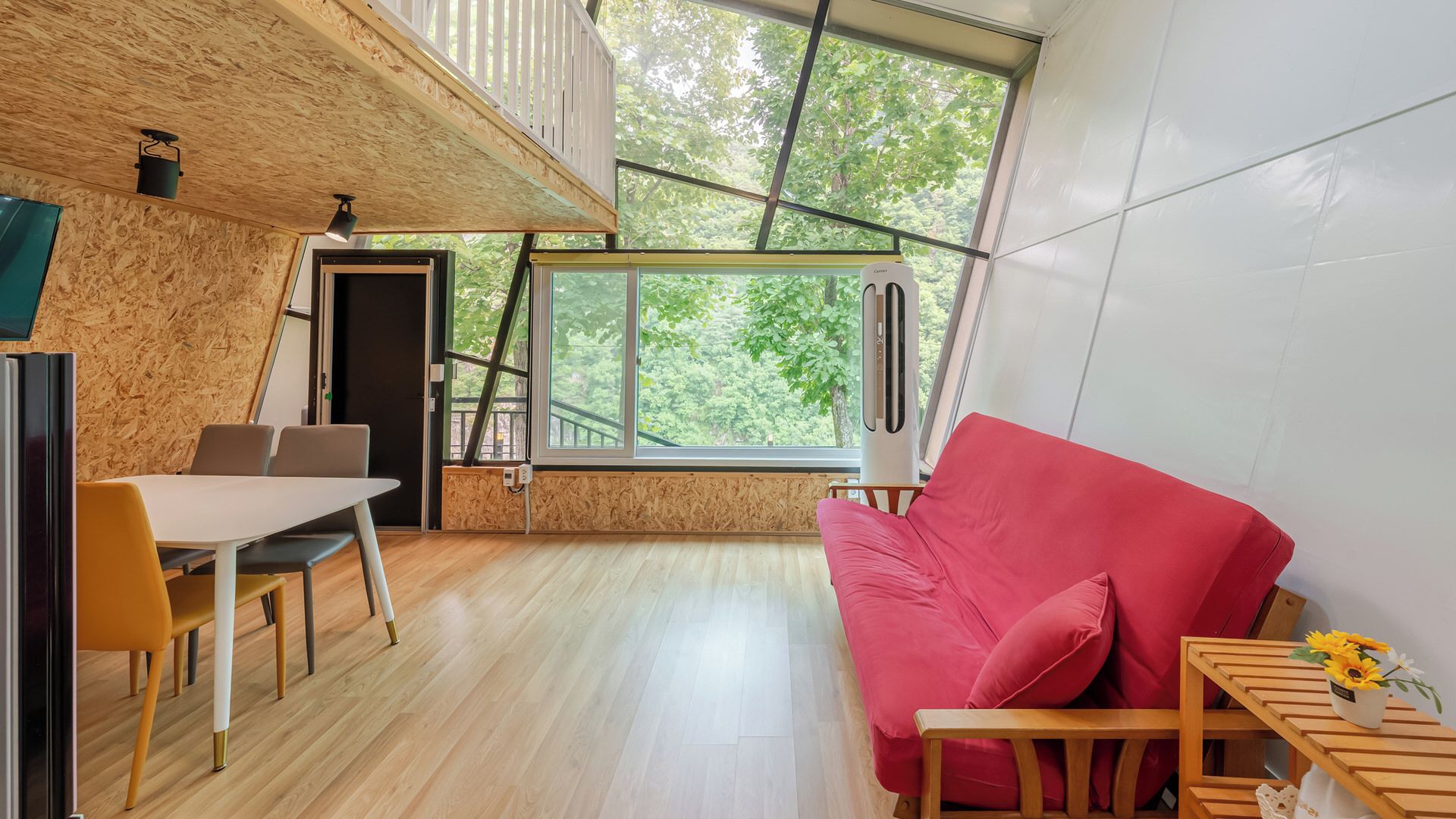 Um interior moderno com piso de madeira, um sofá vermelho, uma mesa de jantar com cadeiras e grandes janelas que oferecem vista para árvores.