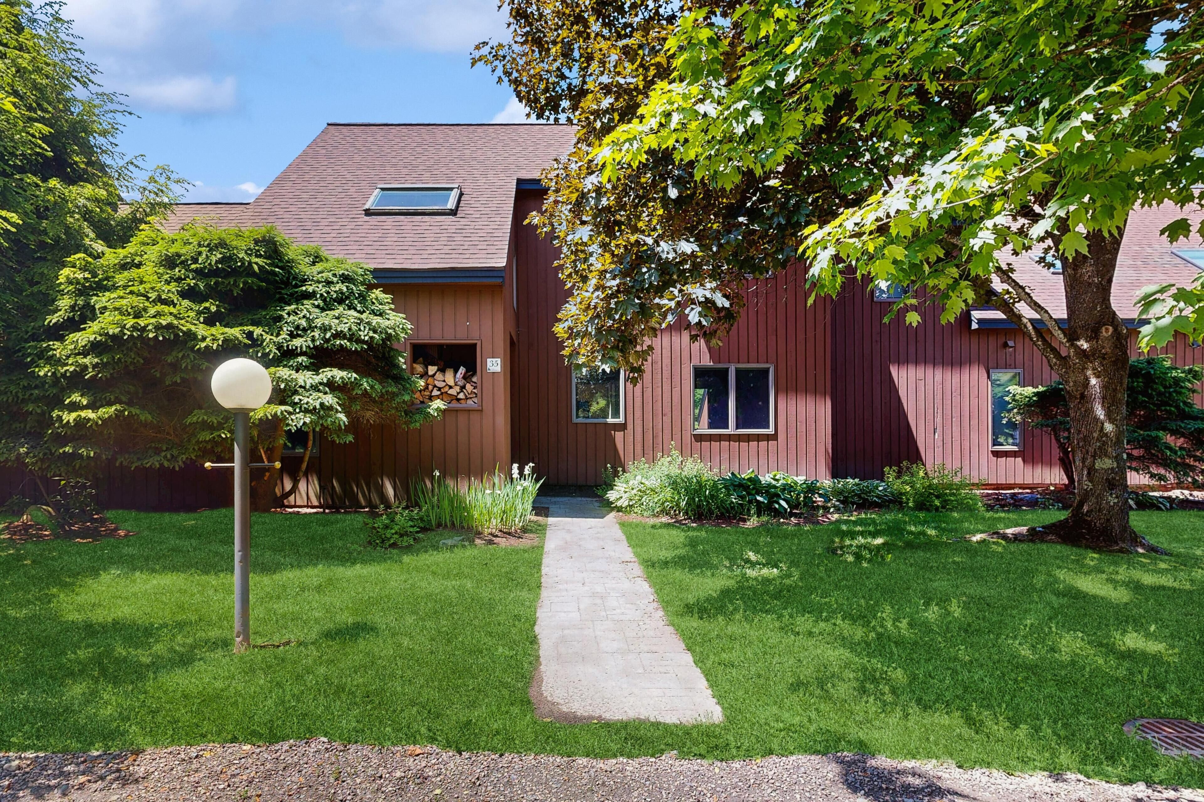 A red building with a paved pathway leading to the entrance, surrounded by greenery and a tree.