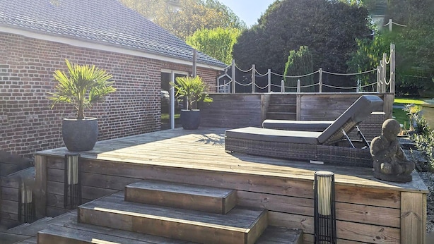Een houten terras met treden naar een zitgedeelte, een standbeeld en een bakstenen gebouw op de achtergrond.