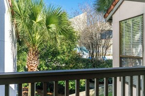 Ein Balkon mit Blick auf eine Palme und andere Bepflanzung, ein weißes Gebäude und ein Fenster mit Rollläden.
