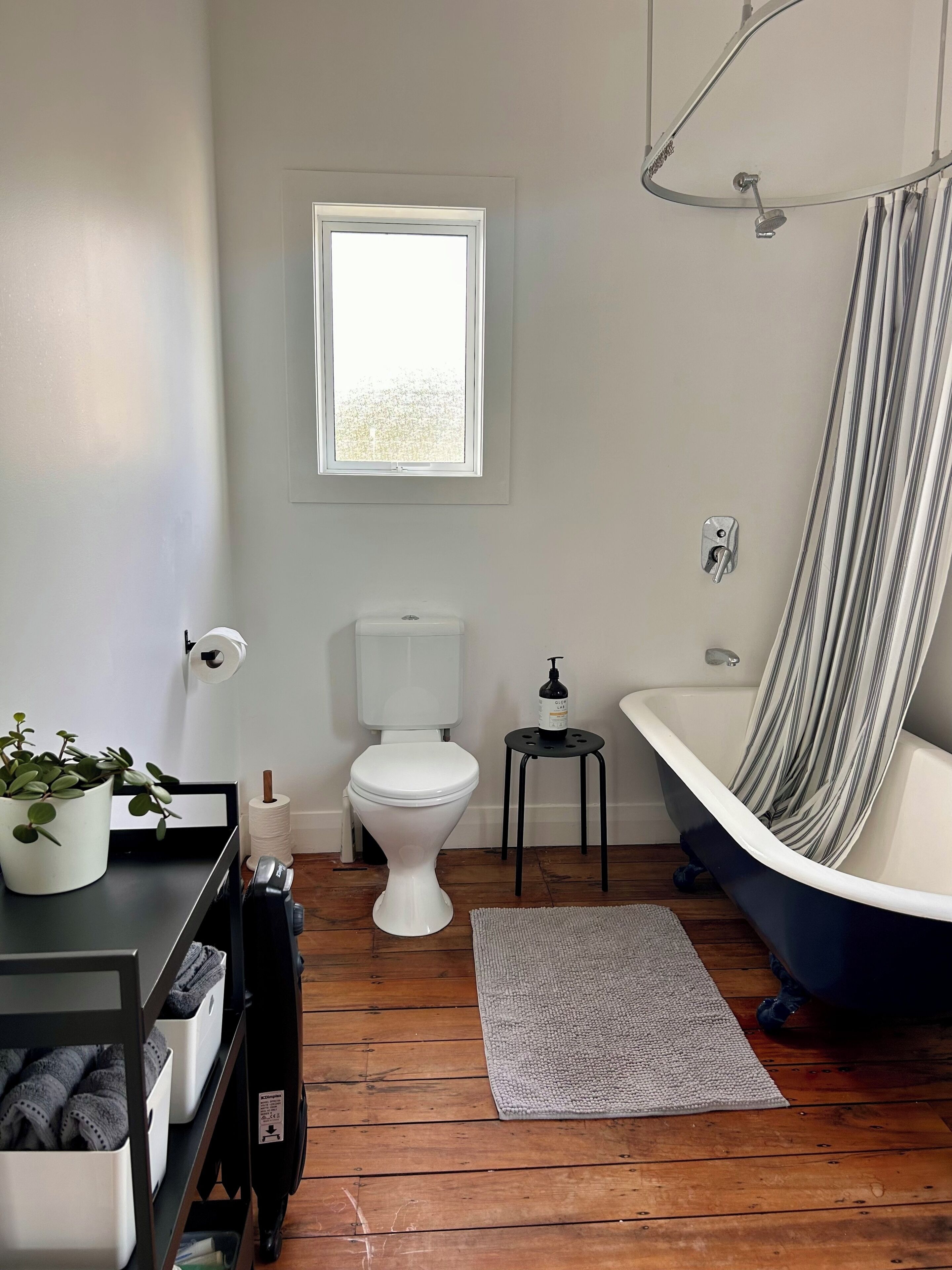 A bathroom with a toilet, a small stool, a bathtub, a window, a shelf with a plant, and a wooden floor.