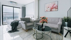 A modern living room with a grey sofa, a round coffee table with a vase of flowers, a large window with a view, and a mounted artwork. - D'. El Dorado Ho Tay Luxury Apartment  (Hanoi)