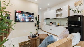 A modern living room with a flat-screen TV, a sofa, a coffee table, and a kitchen area with white cabinets and a black refrigerator. - D'. El Dorado Ho Tay Luxury Apartment  (Hanoi)