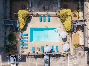 Eine Vogelperspektive auf einen Poolbereich mit Liegestühlen und Sonnenschirmen, umgeben von einem Parkplatz und Grünflächen.
