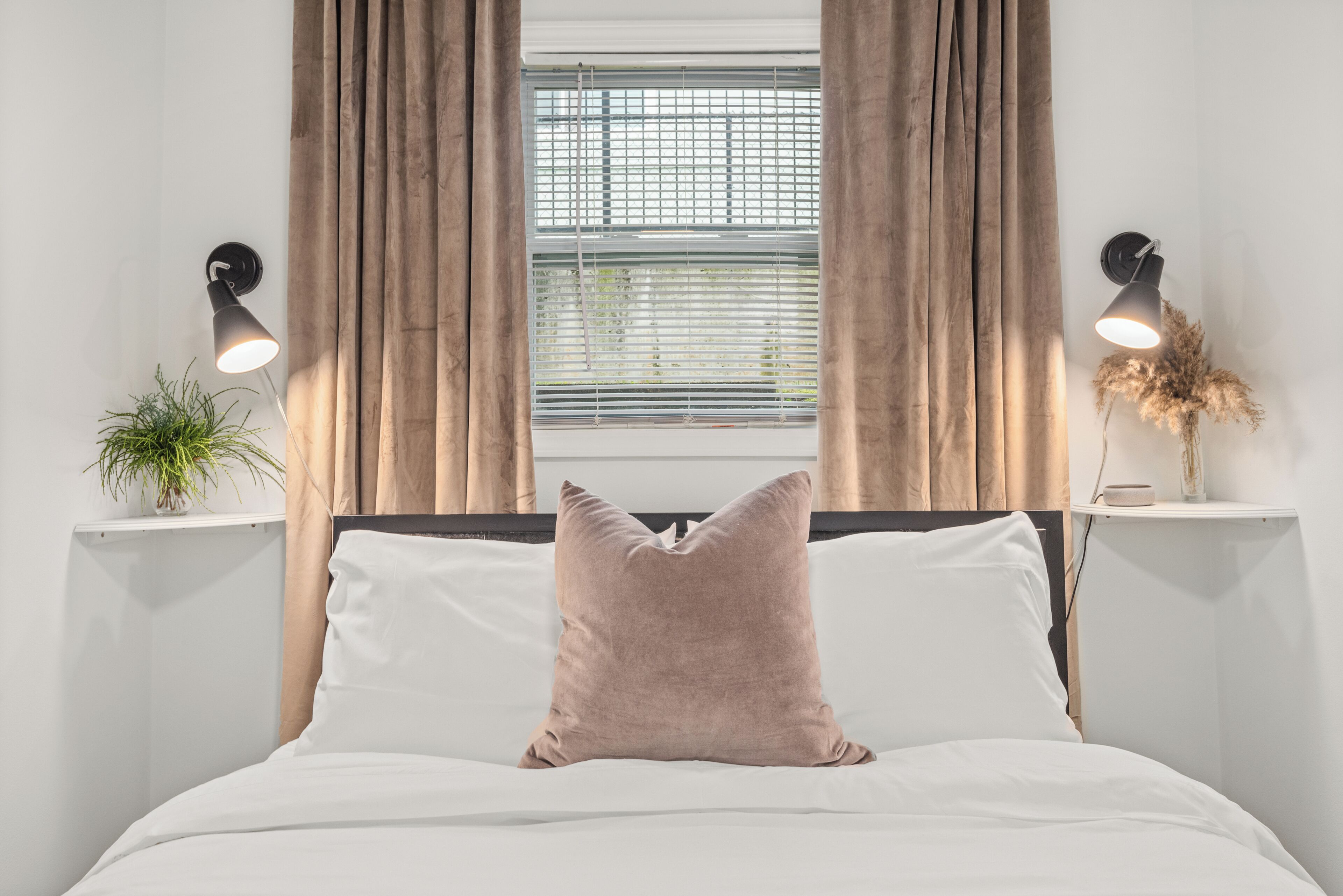 Una cama bien hecha con una única almohada rosa, dos mesitas de noche con lámparas, una ventana con persianas y una planta sobre una de las mesitas.