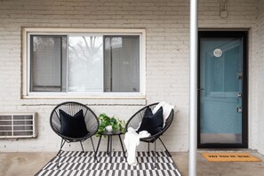 Een kamer met een geruite vloer, twee zwarte stoelen met kussens, een kleine tafel met een plant en een raam met jaloezieën.