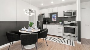 Une cuisine moderne avec des meubles blancs, un réfrigérateur noir, un four à micro-ondes et un four double. On y trouve une table à manger avec des chaises et un luminaire suspendu.