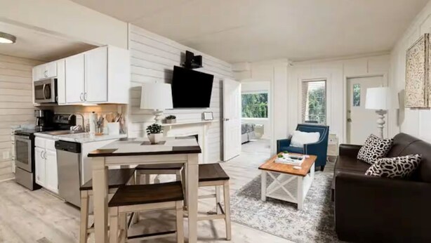 Een moderne keuken met witte kasten, een eethoek met een houten tafel en stoelen, een woonkamer met een bank en salontafel, en een grote televisie aan de muur.