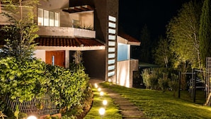 A modern two-story house with a visible balcony, a well-lit pathway, and a garden area.