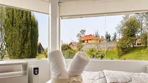 A bedroom with a large window, a bed with a patterned bedspread, a basket with rolled towels, and a radiator.