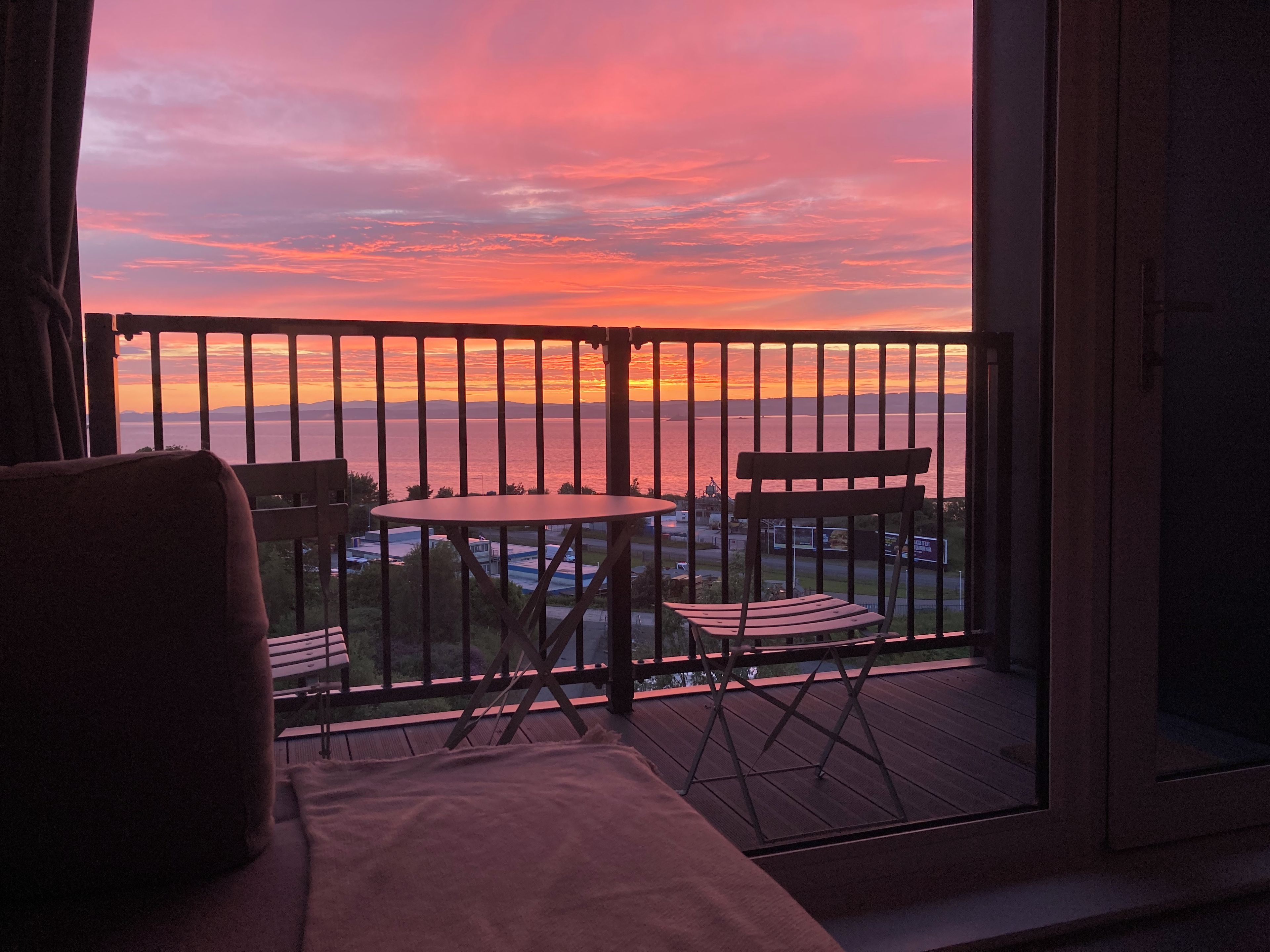 A balcony with a view of the sunset over the sea, a table, two chairs, and a sofa.