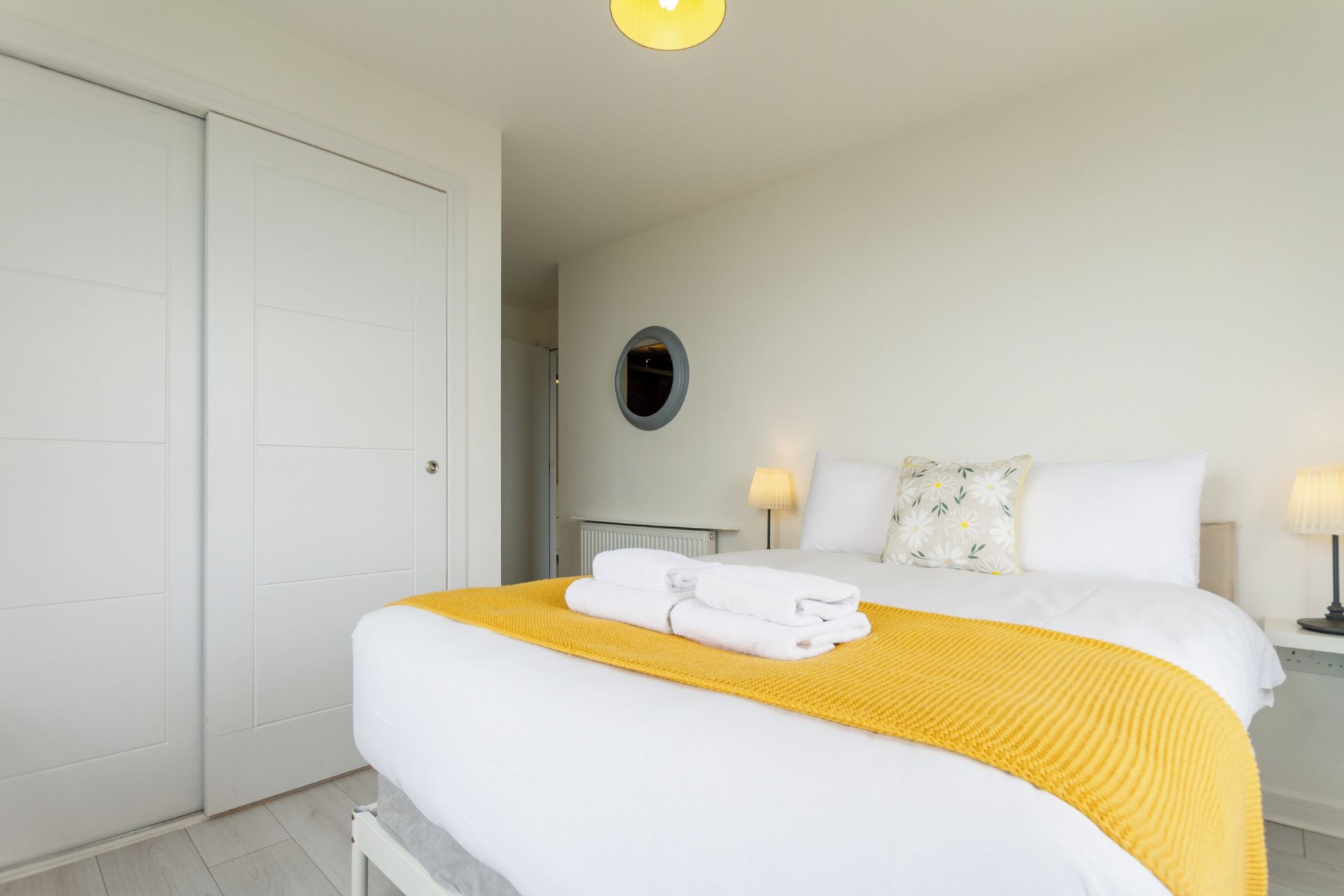 A hotel room with a bed, a bedside table with a lamp, a round mirror, and a white wardrobe.
