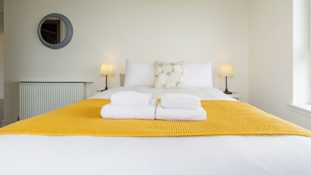 A neatly made bed with white linens and a yellow throw blanket, two bedside lamps, a floral pillow, and a round mirror on the wall. - Lovely Sea View 2BDR Flat in Edinburgh (Edinburgh)