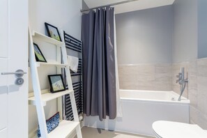 Ein modernes Badezimmer mit Badewanne, Toilette und Duschbereich. Es gibt Regale mit gerahmten Bildern und einen Handtuchhalter.