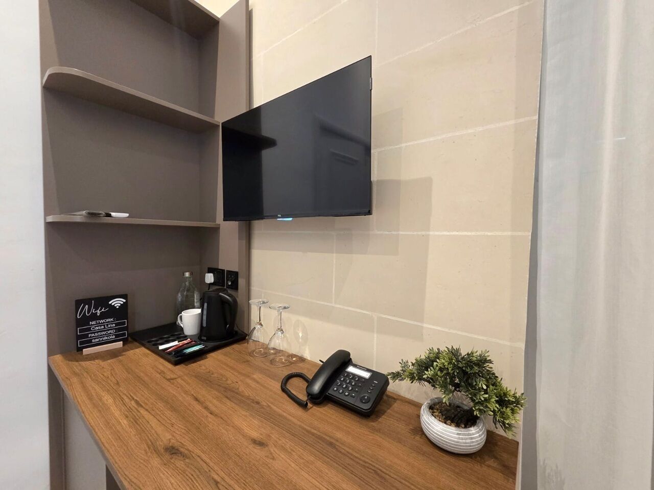 Un espace d’accueil d’hôtel avec un comptoir en bois, un téléviseur à écran plat fixé au mur, un téléphone, une machine à café, un panneau indiquant les informations relatives au Wi-Fi et une petite plante en pot.