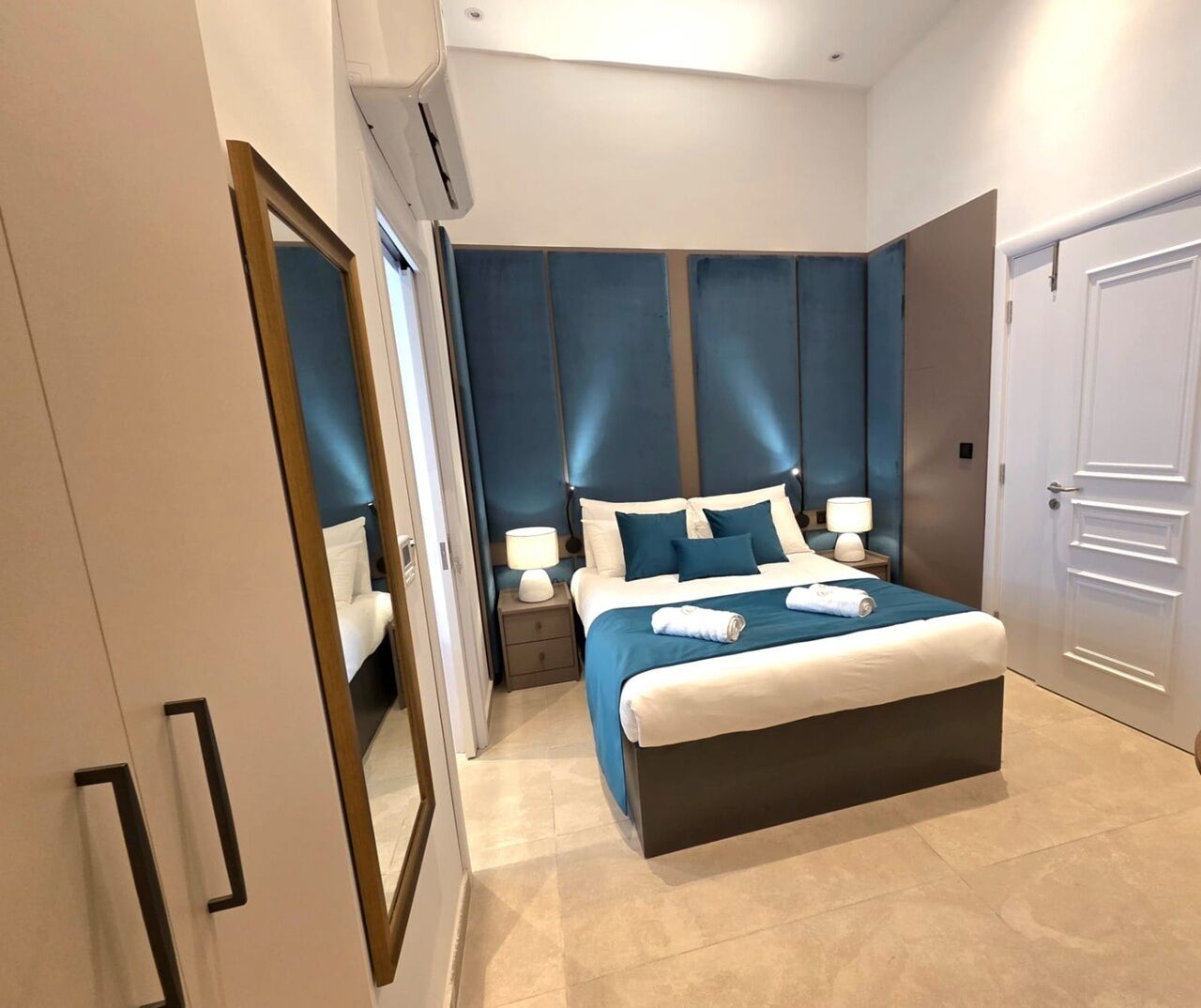 A modern hotel bedroom with a large bed, bedside tables, a mirror, and a wardrobe.