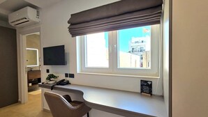 Quarto com escrivaninha, cadeira, janela com cortinas, ar condicionado e televisão de tela plana.