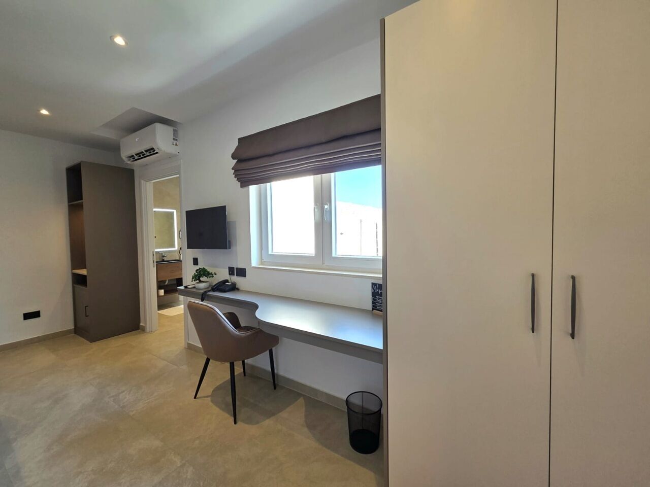 Ein modernes Zimmer mit Schreibtisch, Stuhl, Fenster und Kleiderschrank.