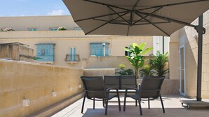 Um pátio externo com mesa, cadeiras, um grande guarda-sol e plantas em vasos.