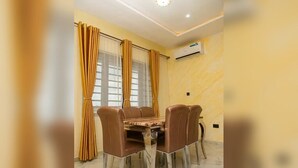 A dining area with a table and chairs, a window with curtains, and an air conditioning unit on the wall. - City-Chic Ikeja 2BR Escape: 24/7 Power, Fast WiFi, Airport, Shops & Eats - Lagos (Ikeja)