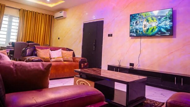 A modern living room with a sofa, a coffee table, and a flat-screen TV mounted on the wall. - City-Chic Ikeja 2BR Escape: 24/7 Power, Fast WiFi, Airport, Shops & Eats - Lagos (Ikeja)