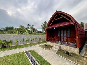 A wooden cabin with a sloped roof, a porch with a railing, and a stone wall in the background. - Kasturi Villas Bedugul by AGATA (Tabanan)