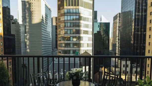 Ein Balkon mit rundem Tisch und Stühlen, Blumentöpfen und Blick auf hohe Gebäude.