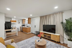 A modern living room with a flat-screen TV, a dining area with a kitchen in the background, and a large window with curtains.