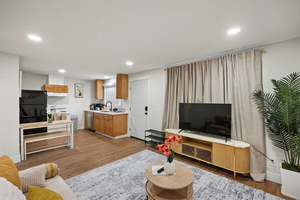 A modern living room with a flat-screen TV, a dining area with a kitchen in the background, and a large window with curtains.