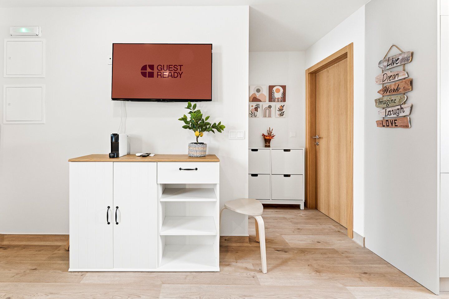 A modern hotel room with a white wardrobe, a small desk with a plant, a television displaying 'GUEST READY', and a wooden door.