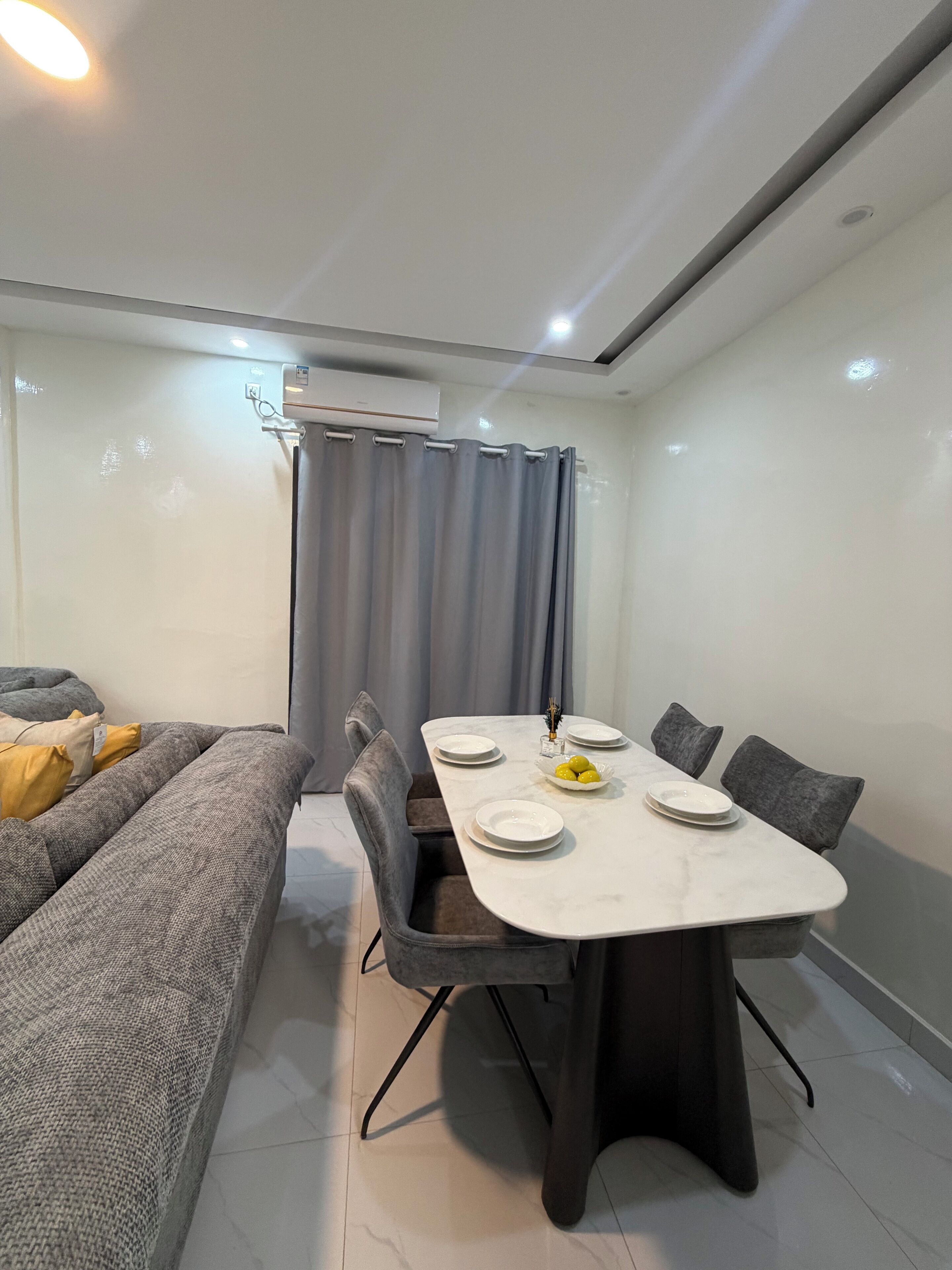 Un espace repas moderne avec une table en marbre, des chaises recouvertes d’un tissu gris et un canapé gris agrémenté de coussins jaunes.