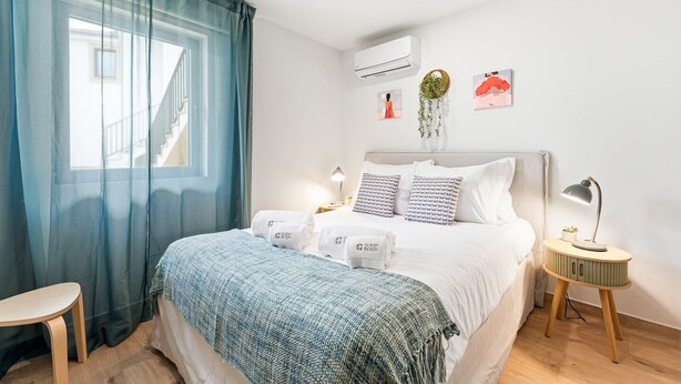 Ein ordentlich eingerichtetes Schlafzimmer mit Bett, Nachttischen, einem Fenster mit Vorhängen, einer Klimaanlage und Wanddekorationen.