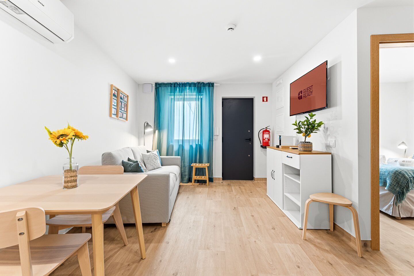 A modern living room with a sofa, a dining table with chairs, a small kitchen area, and a TV mounted on the wall.