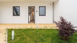 Ein modernes, zweistöckiges Gebäude mit Eingang, einem Balkon mit Sitzgelegenheit und einem kleinen Gartenbereich mit einem Strauch.