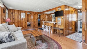 Una habitación con sofá, mesa de centro, televisor, barra con taburetes y decoración de madera.