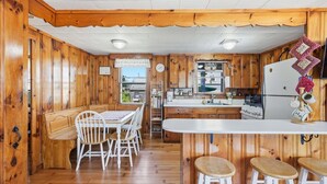 Cocina estilo cabaña con paredes de madera, comedor con sillas blancas, barra con taburetes y vistas al exterior.